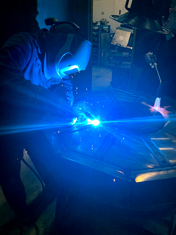 Welder fabricating a component, arc light illuminating the workshop at CNTL